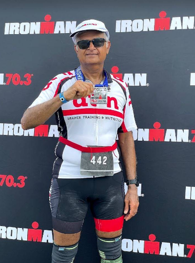 Sanjay Shah stands in front of an Iron Man 70.3 poster after a race and holds a medal for completing the triathlon